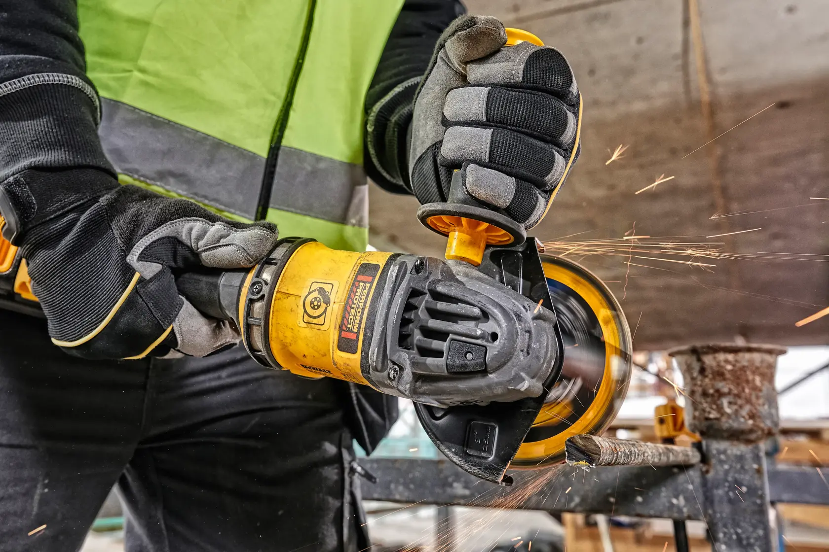 Worker uses yellow DEWALT angle grinder to cut metal, wearing gloves and safety vest. Sparks visible during operation.