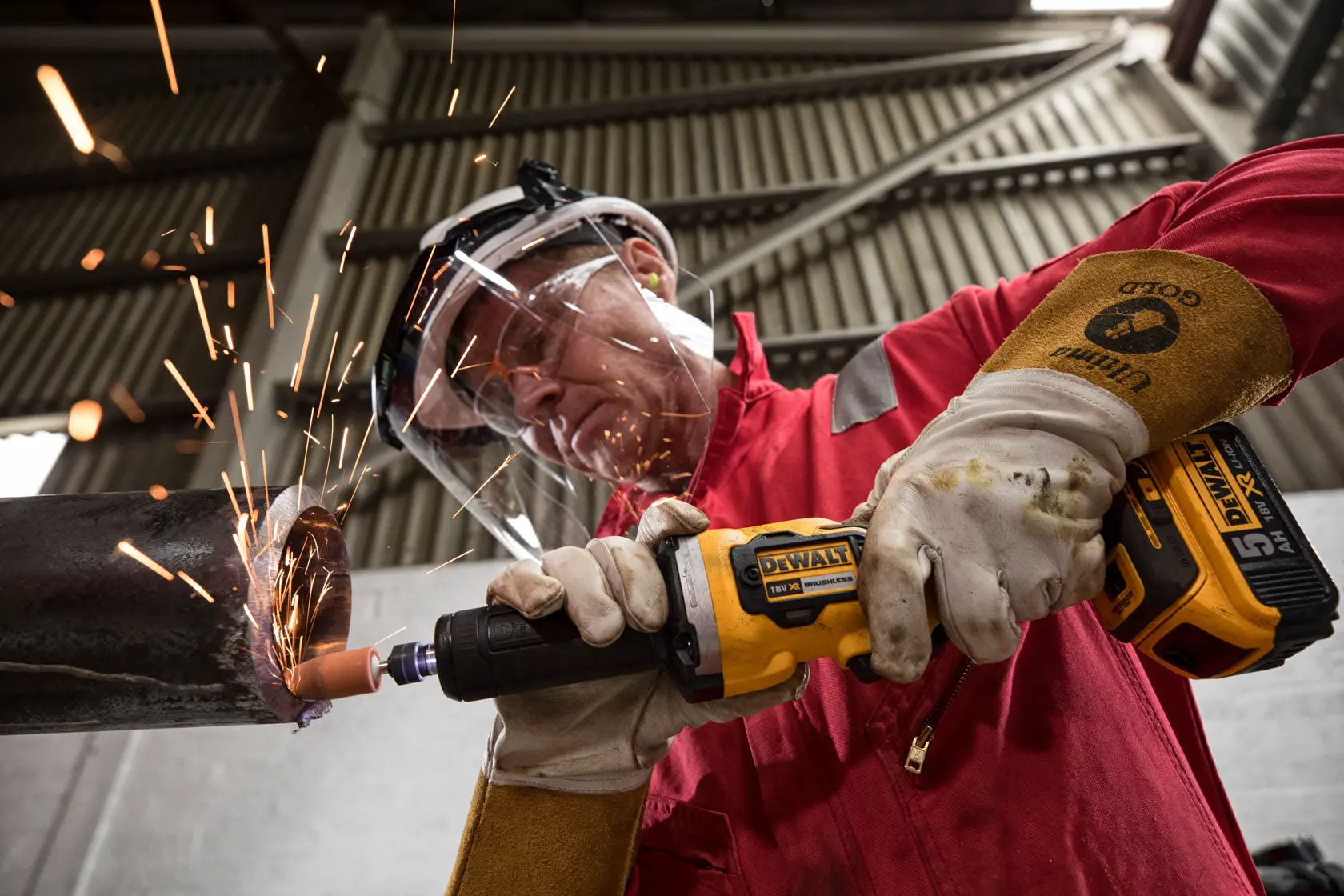 En person med beskyttelseshansker og sikkerhetsutstyr bruker en DEWALT DCG426N batteridrevet slipemaskin på et metallrør, og det oppstår gnister i et industrielt miljø.