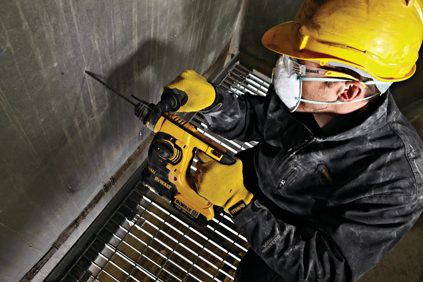 Worker uses a DEWALT DCH253N rotary hammer drill on a concrete wall, wearing safety gear and gloves.