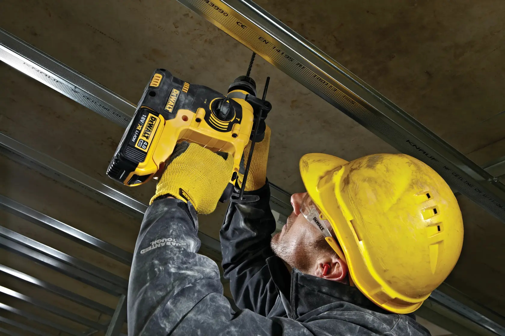 Person in safety gear uses a DEWALT DCH253N power tool to drill into a ceiling structure at a worksite.