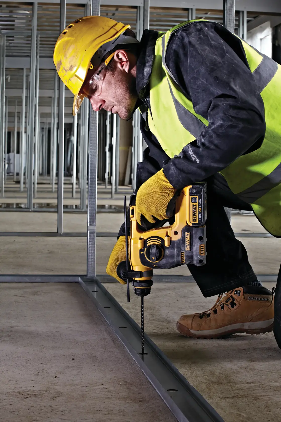 A construction worker wearing a yellow hard hat, safety vest, gloves, and boots is using a DEWALT DCH253 cordless rotary hammer drill to drill into a metal frame on a construction site.