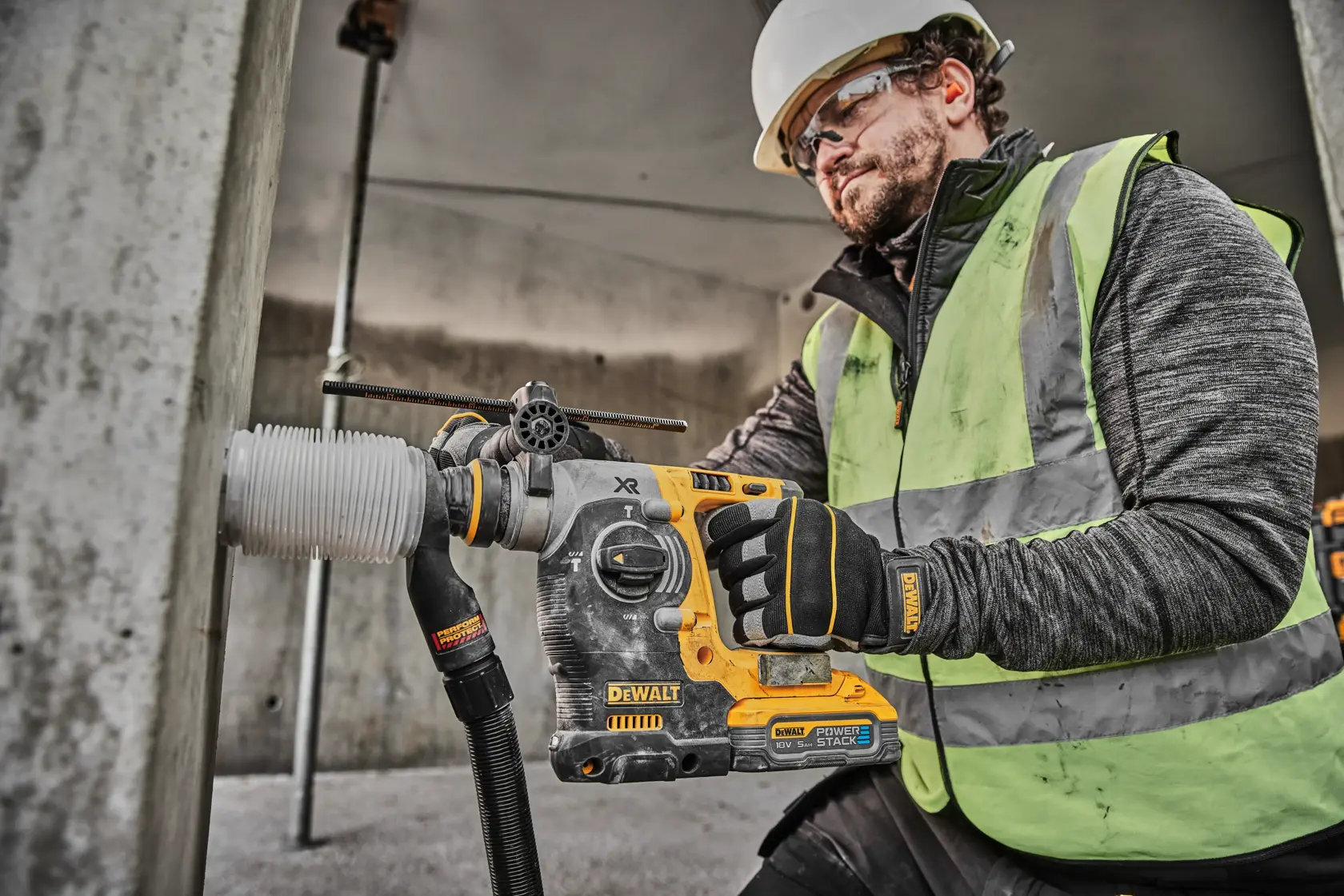 Un operaio edile con casco di sicurezza e giubbotto ad alta visibilità utilizza un martello rotativo a batteria DEWALT DCH273H2T per forare una colonna di cemento. L'operaio indossa anche guanti DEWALT.