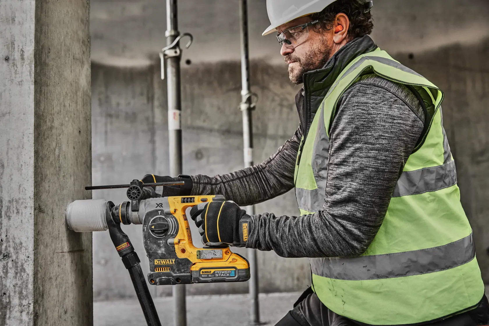 Una persona con gilet di sicurezza e guanti usa un martello rotante a batteria DEWALT DCH273H2T per forare un pilastro di cemento in un cantiere. Il volto dell'utente è sfocato per la privacy.