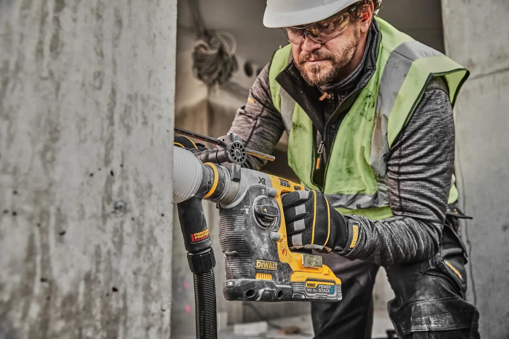 Un operaio edile utilizza un martello rotante DEWALT DCH273H2T con aspirazione della polvere per forare il cemento. L'operaio indossa guanti di sicurezza, un gilet ad alta visibilità e un casco. Il trapano è della marca DEWALT con batteria PowerStack.