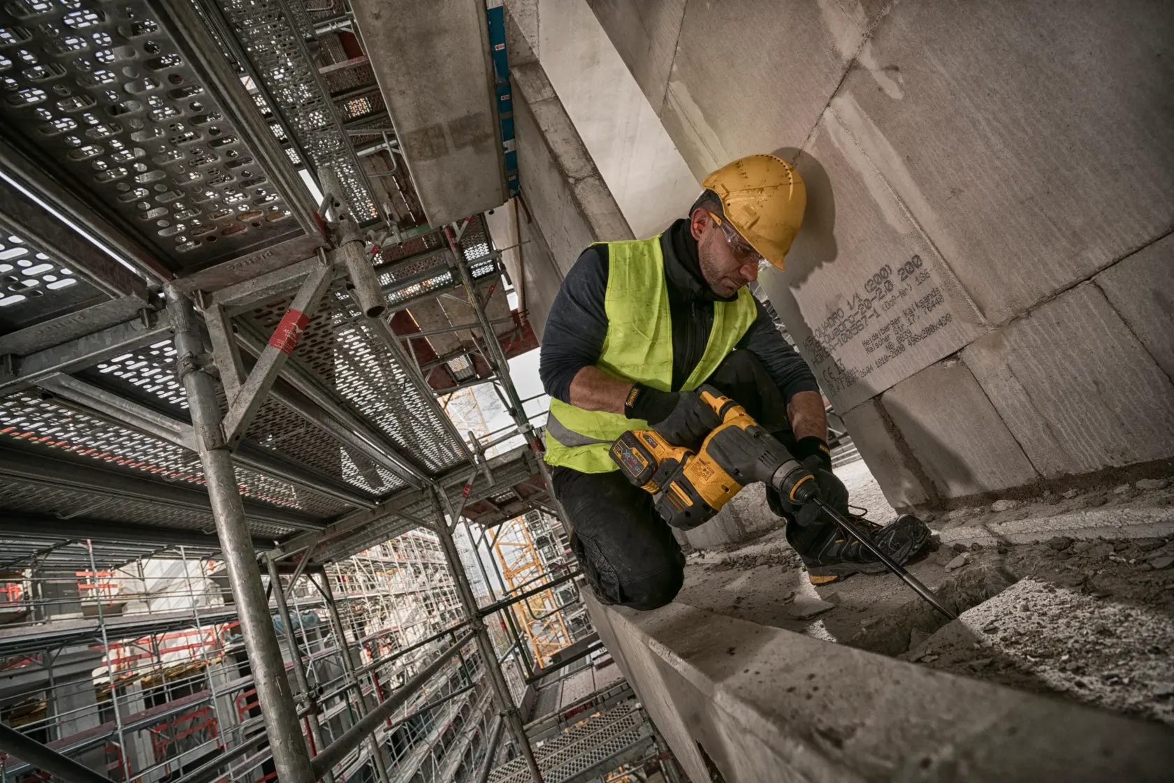 Worker using a DEWALT rotary hammer drill on a construction site with scaffolding in the background.