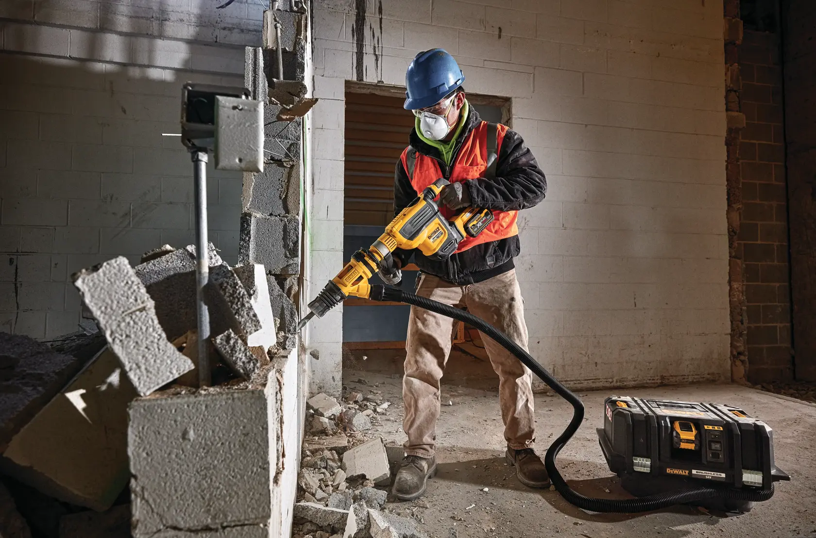 Worker uses DEWALT demolition tool to break a concrete wall, wearing safety gear and mask on a construction site.