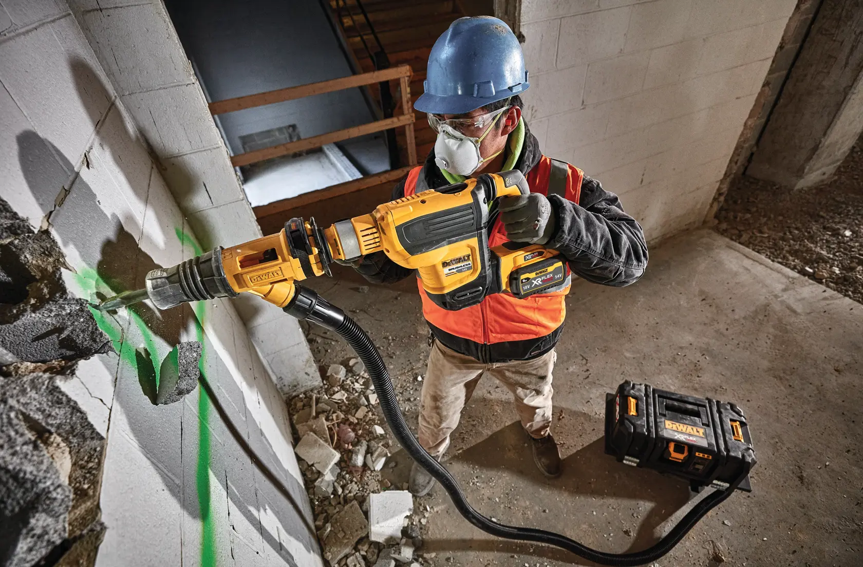 Worker uses a DEWALT demolition tool to break concrete wall indoors, wearing safety gear and helmet.