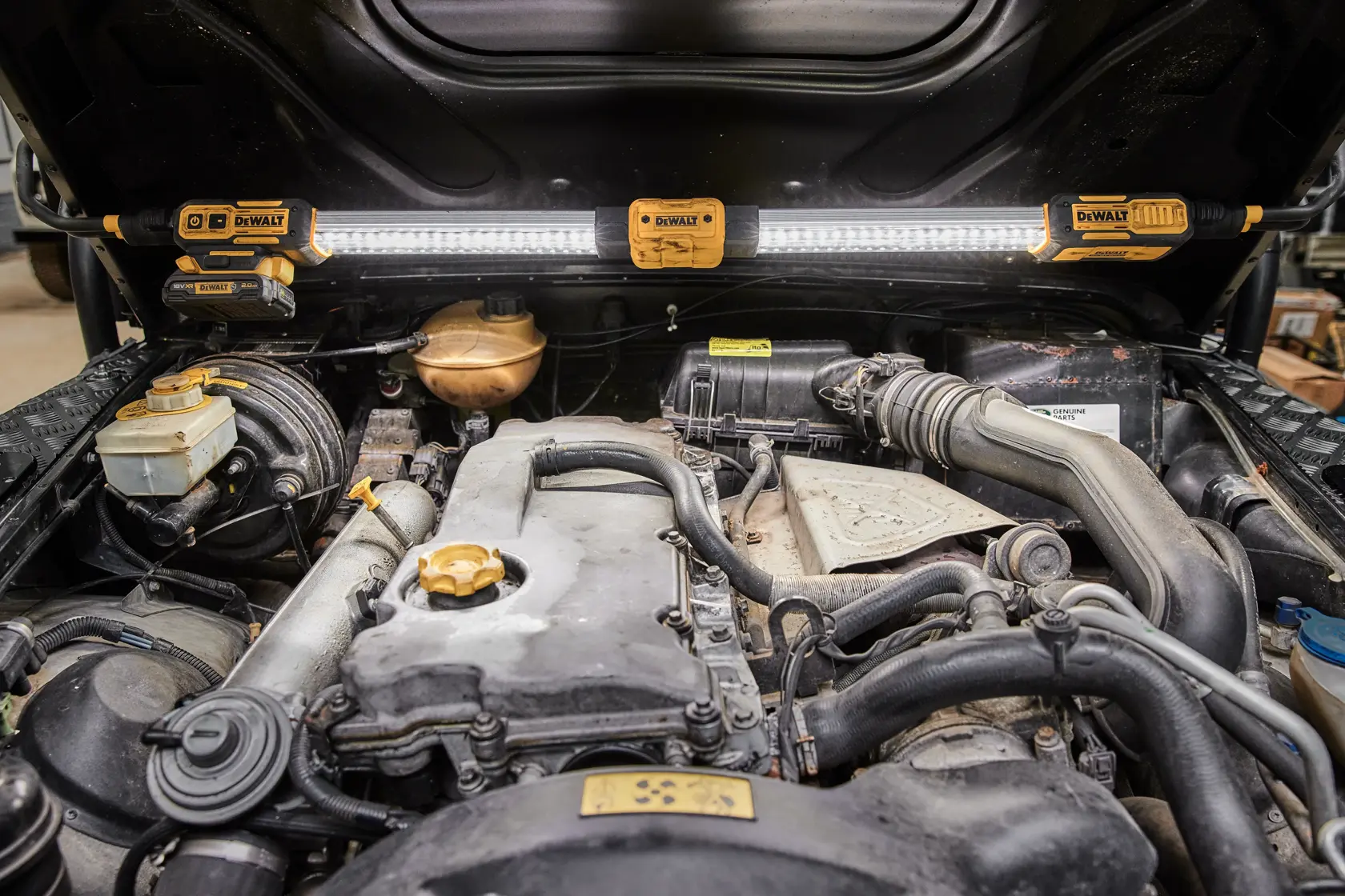 A DEWALT DCL045 LED work light is mounted under the open hood of a vehicle, illuminating the engine compartment for better visibility while working.