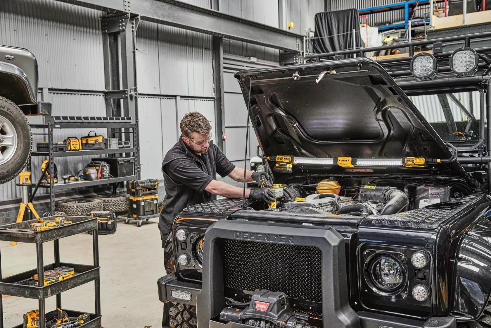 A person is working on the engine of a black Land Rover Defender inside a workshop. There are DEWALT tools visible on the shelves and next to the vehicle, and the DEWALT DCL045 work light is positioned under the open hood to illuminate the engine area.