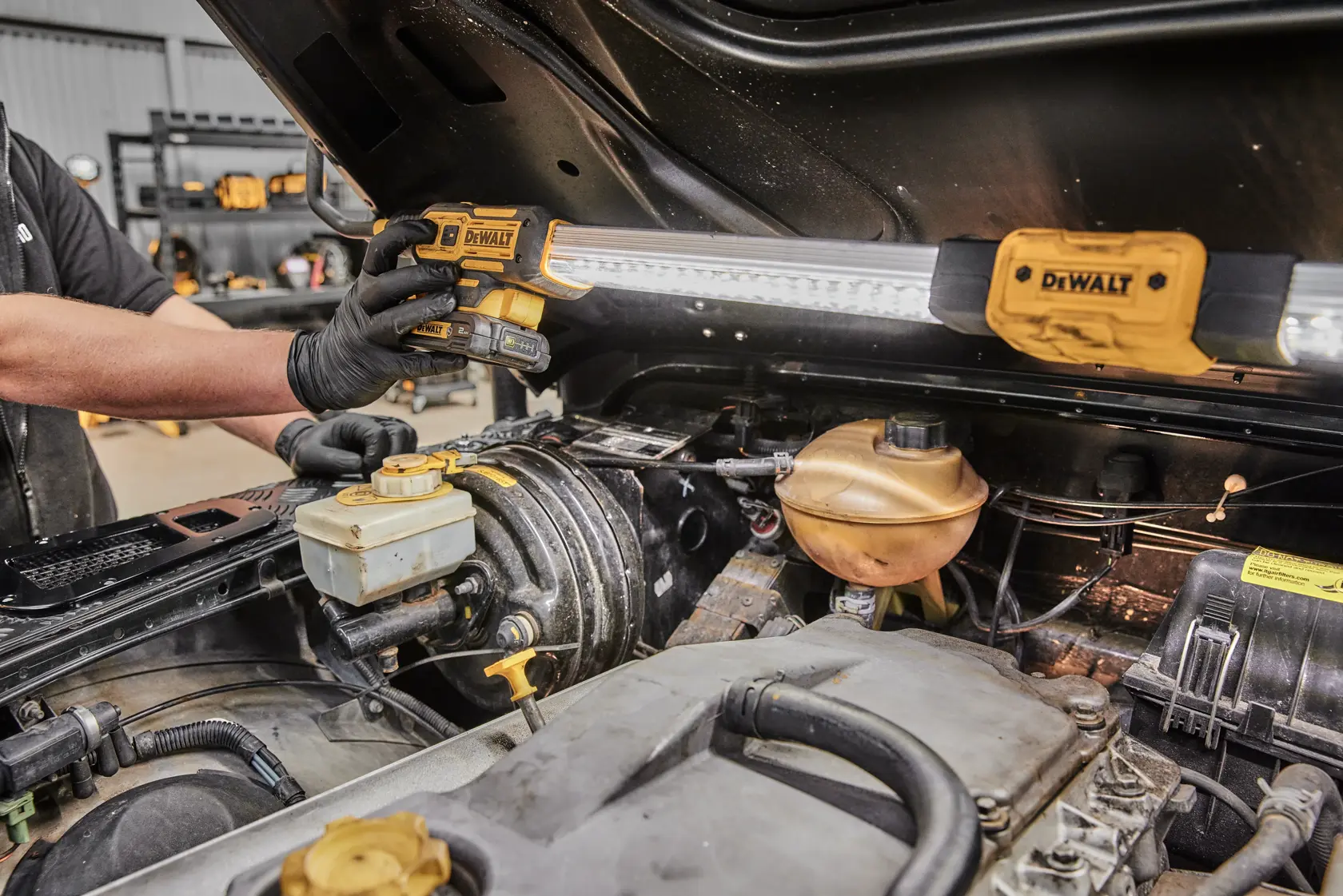 A mechanic uses a DEWALT DCL045 work light to illuminate the engine bay of a vehicle. The work light is mounted under the raised hood to provide bright lighting for maintenance and repair tasks.
