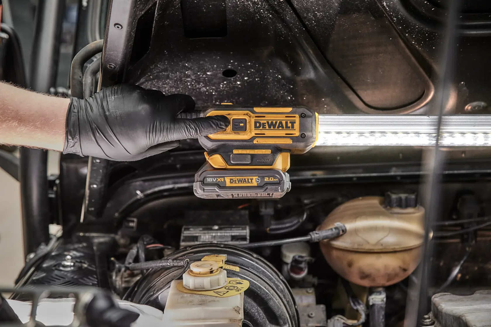 A gloved hand is holding a DEWALT DCL045 cordless LED work light under the hood of a vehicle, illuminating the engine area.