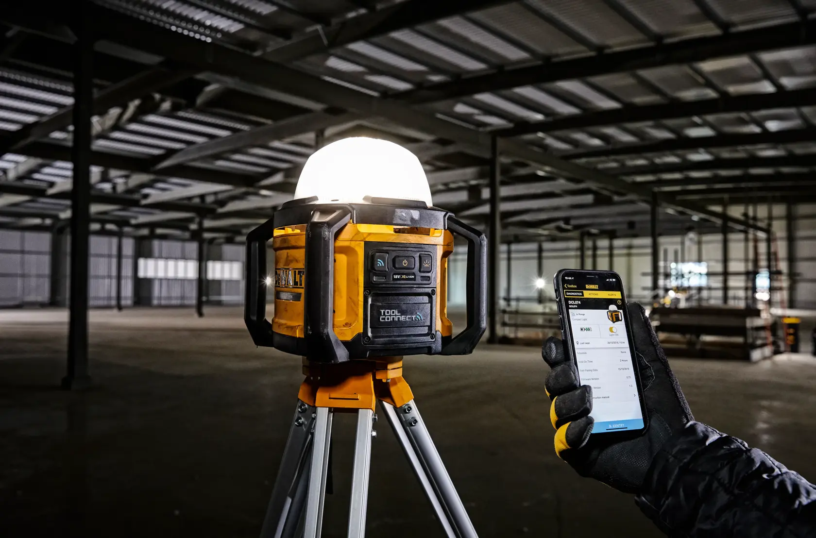 DEWALT work light on tripod in warehouse, controlled by a gloved hand using a smartphone.