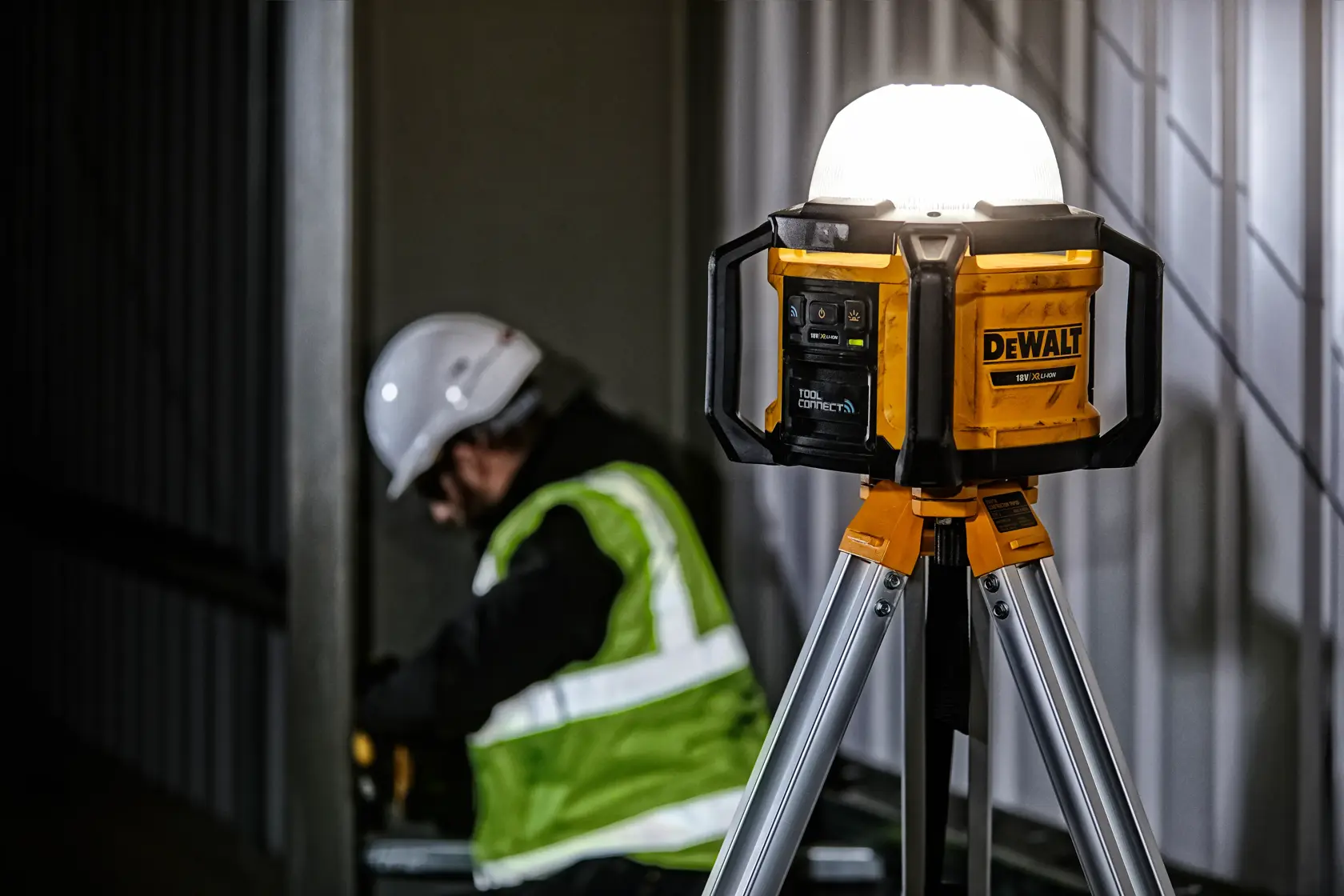 DEWALT DCL074 LED work light on tripod, used at construction site with worker in safety gear in background.