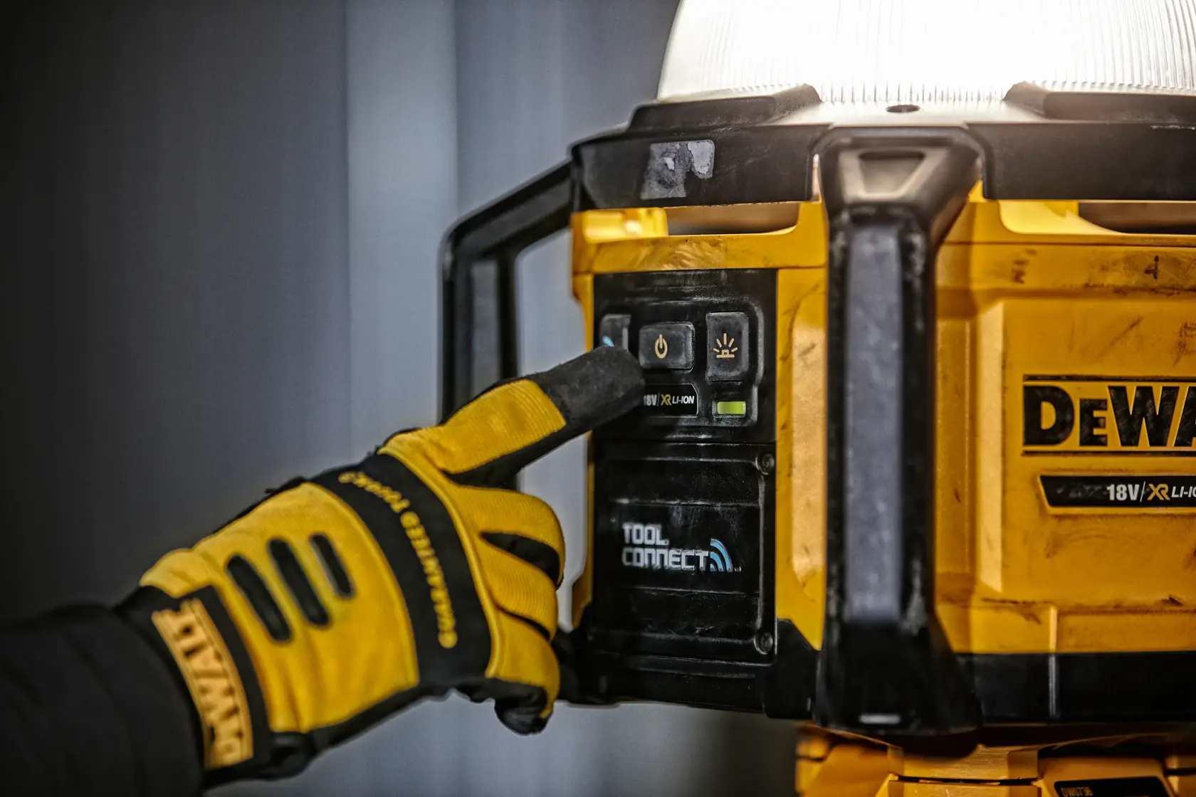 Gloved hand presses button on yellow DEWALT DCL074 work light with Tool Connect feature.