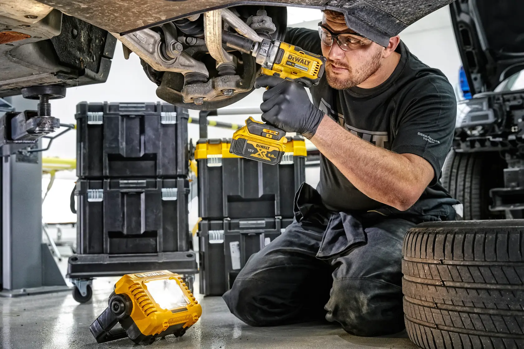 Mechanic uses DEWALT tools and LED work light under a car, with tool storage boxes visible in the background.