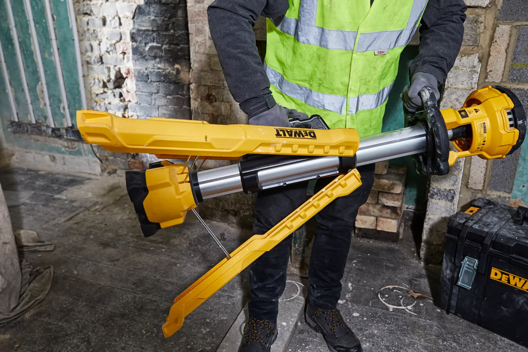 Person sets up a yellow DEWALT tripod work light in an industrial environment with brick walls and equipment.