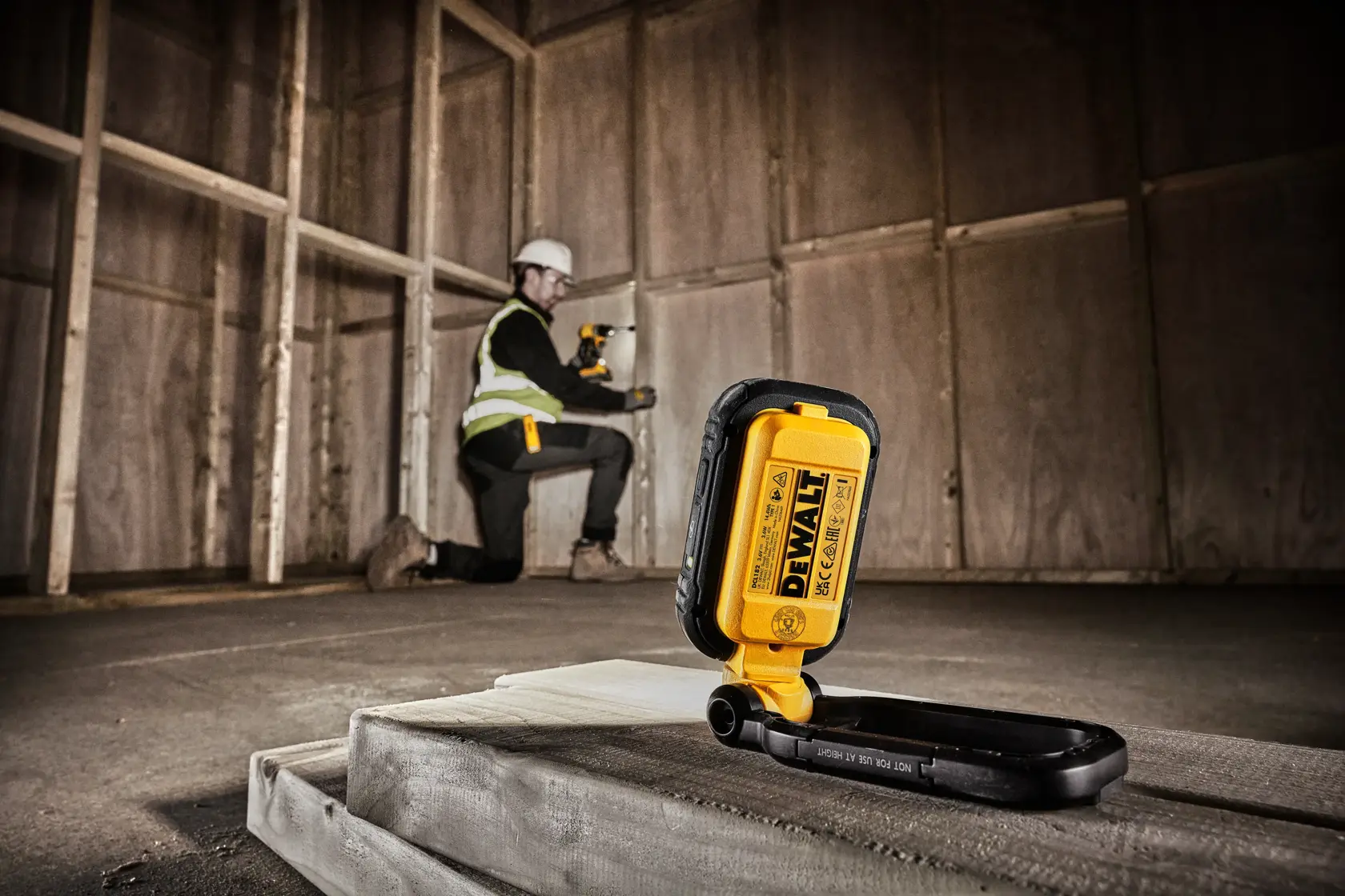 En DEWALT DCL182 bærbar arbeidslampe er plassert på en betongflate i et uferdig rom med treramme. I bakgrunnen kneler en person med sikkerhetsvest og bruker et trådløst verktøy på veggen.