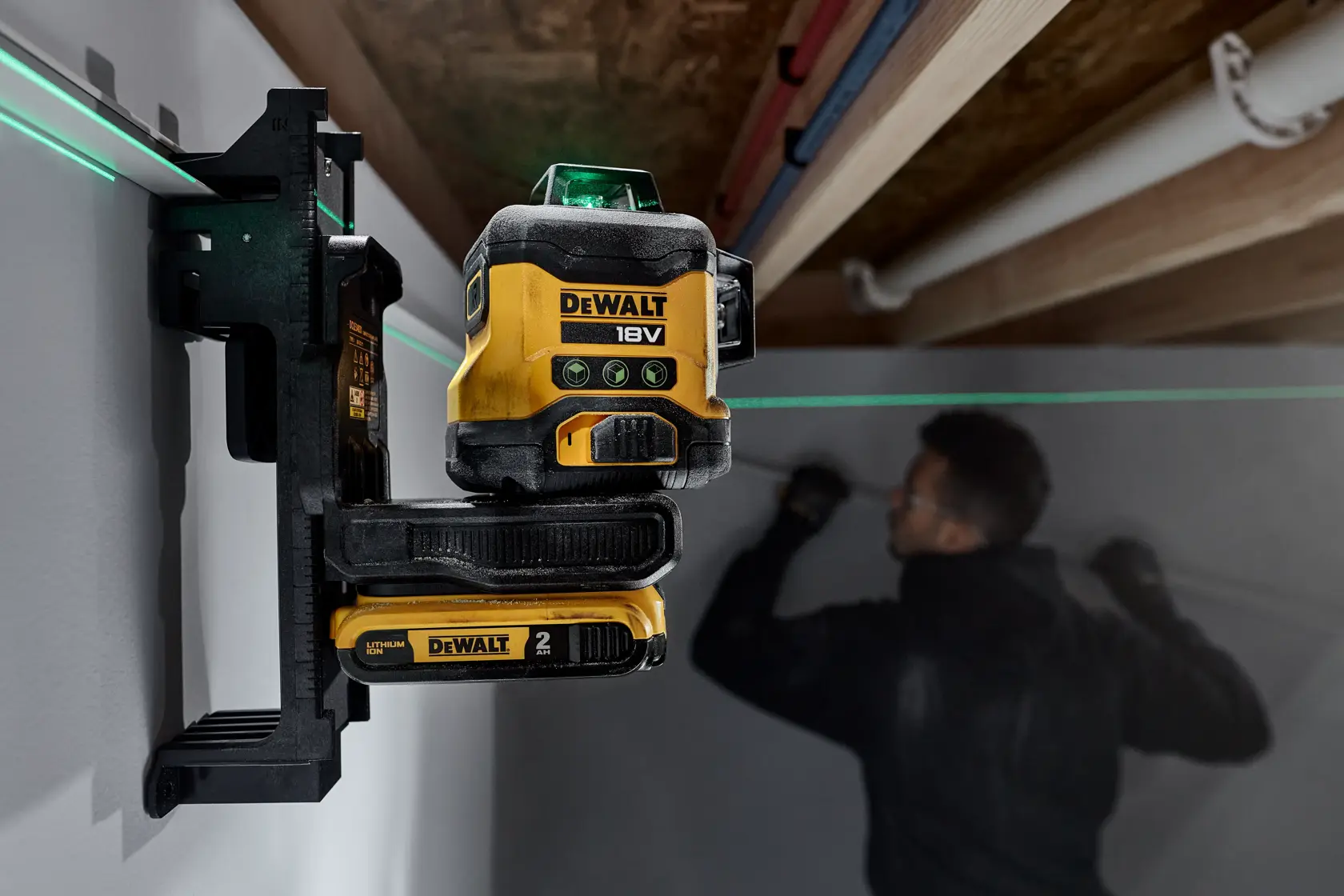 DEWALT 18V cross line laser level mounted on a wall, projecting a green laser line across a room where a person is working on the wall in the background.