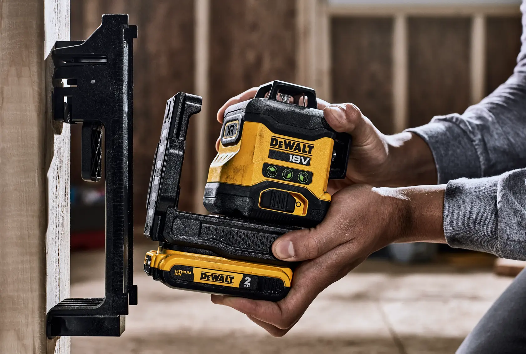 A person is holding a DEWALT 18V laser level with a 2 Ah battery, positioning it next to a wall-mounted bracket in an indoor construction setting.