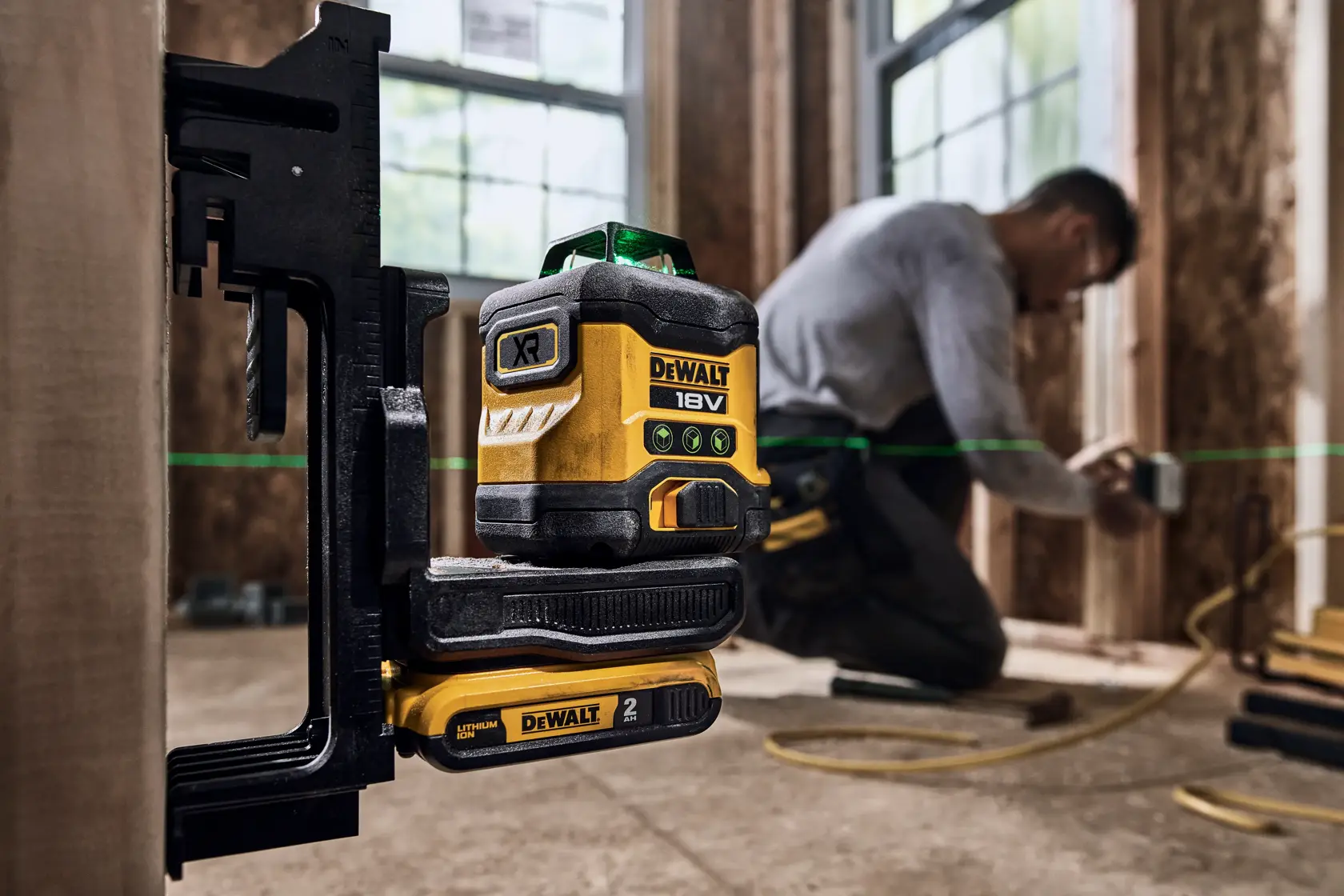 DEWALT 18V XR green line laser level mounted on a bracket with battery attached, being used on a construction site. A person is working in the background with blurred face, and the green laser line is visible across the workspace.