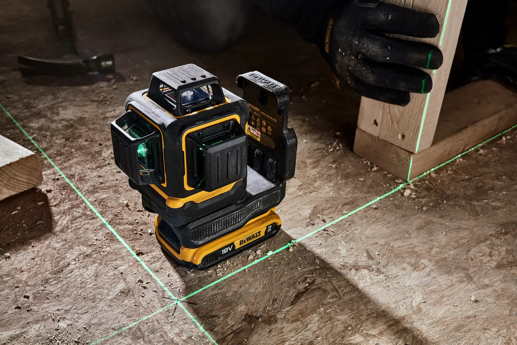 A DEWALT DCLE34031N 18V laser level device projects green laser lines on a construction surface. A gloved hand holds a wooden beam, aligning it with the laser lines. Construction tools and materials are visible in the background.