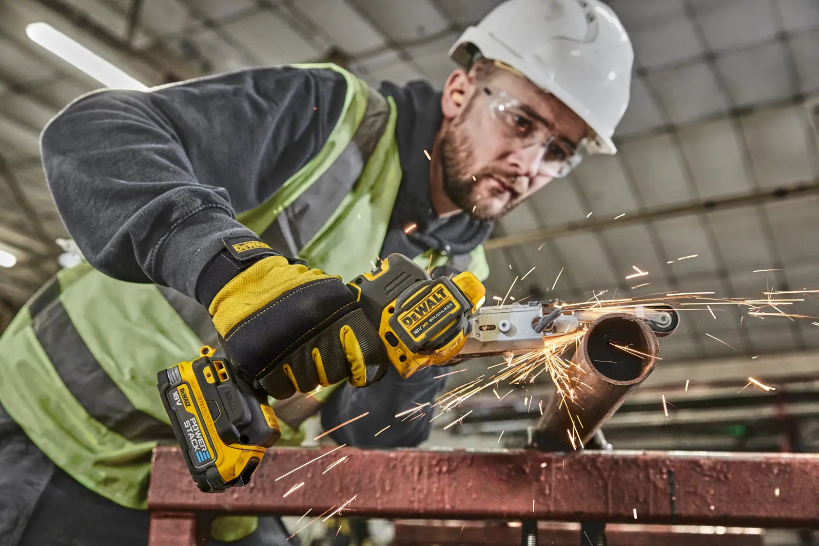 Worker uses DEWALT cordless tool to cut metal pipe, producing sparks. Indoor industrial setting, safety gear visible.