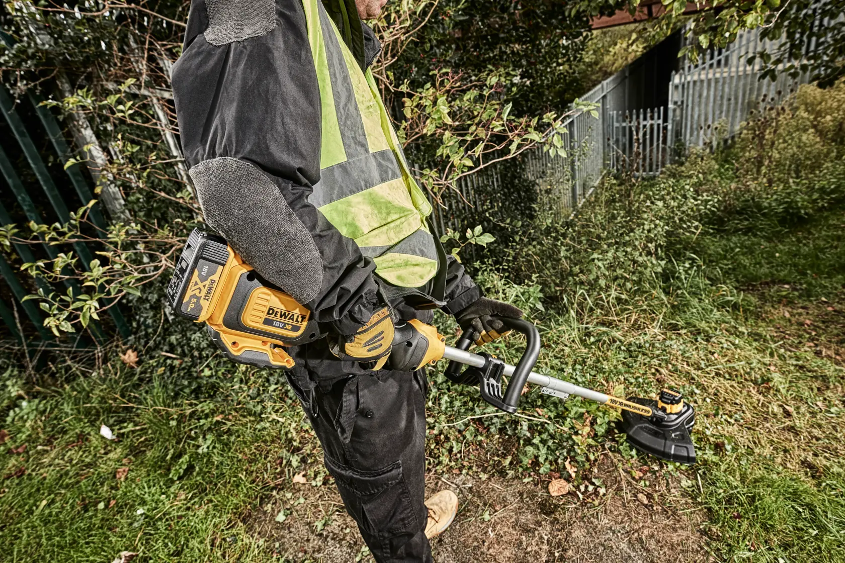 Persona usa un decespugliatore DEWALT DCM561PBS all’aperto, taglia erba vicino a una recinzione.