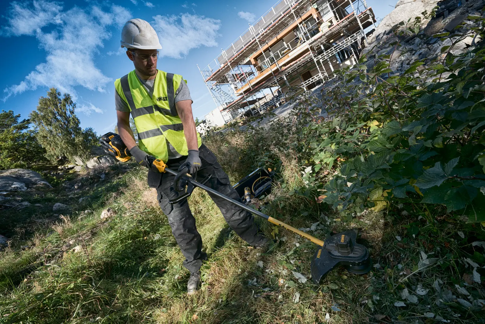 Worker uses DEWALT brush cutter outdoors, wearing safety gear near a construction site with scaffolding.