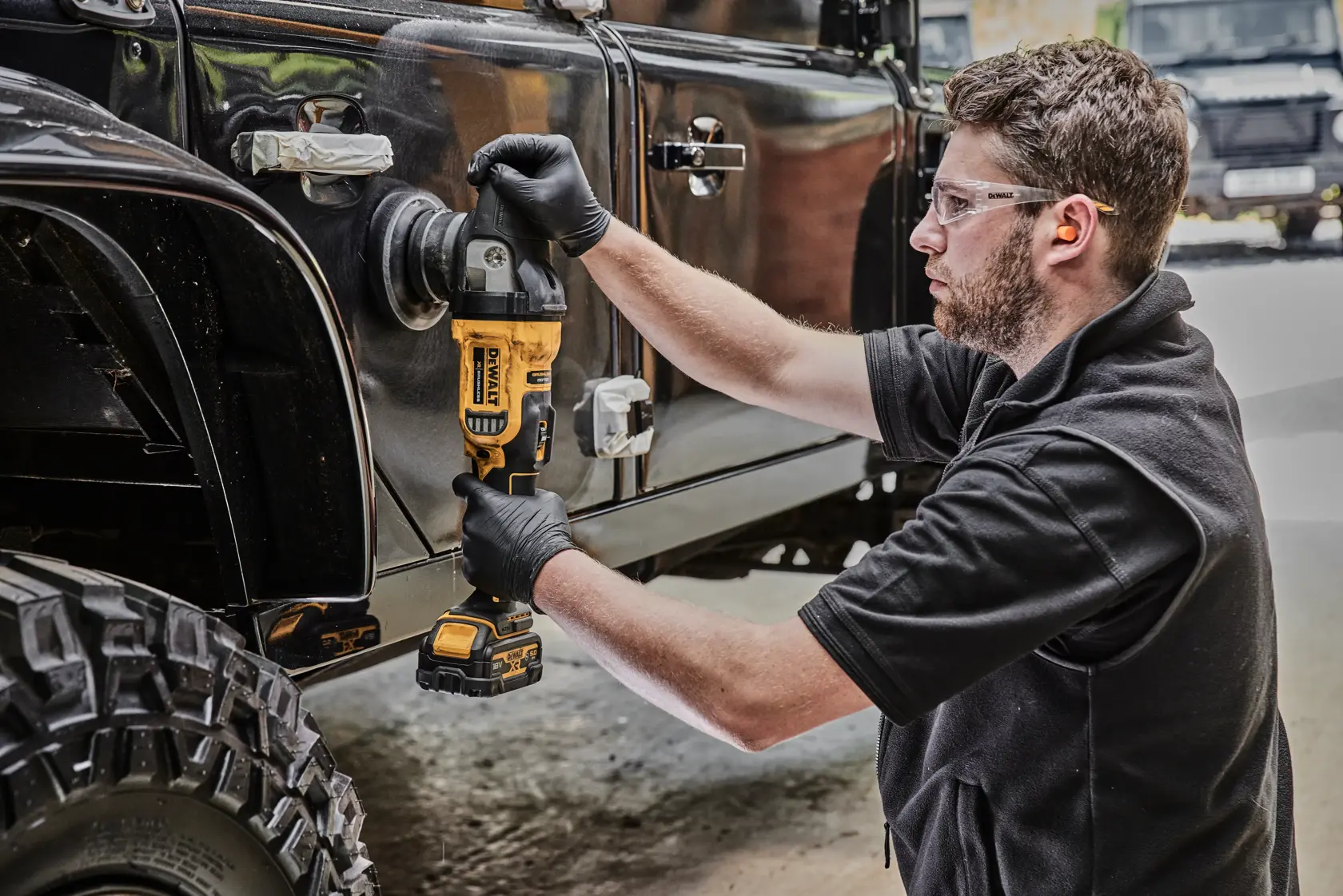 A person wearing black gloves uses a DEWALT cordless polisher (model DCM848N) to polish the side of a black off-road vehicle. The scene takes place in a workshop or garage.