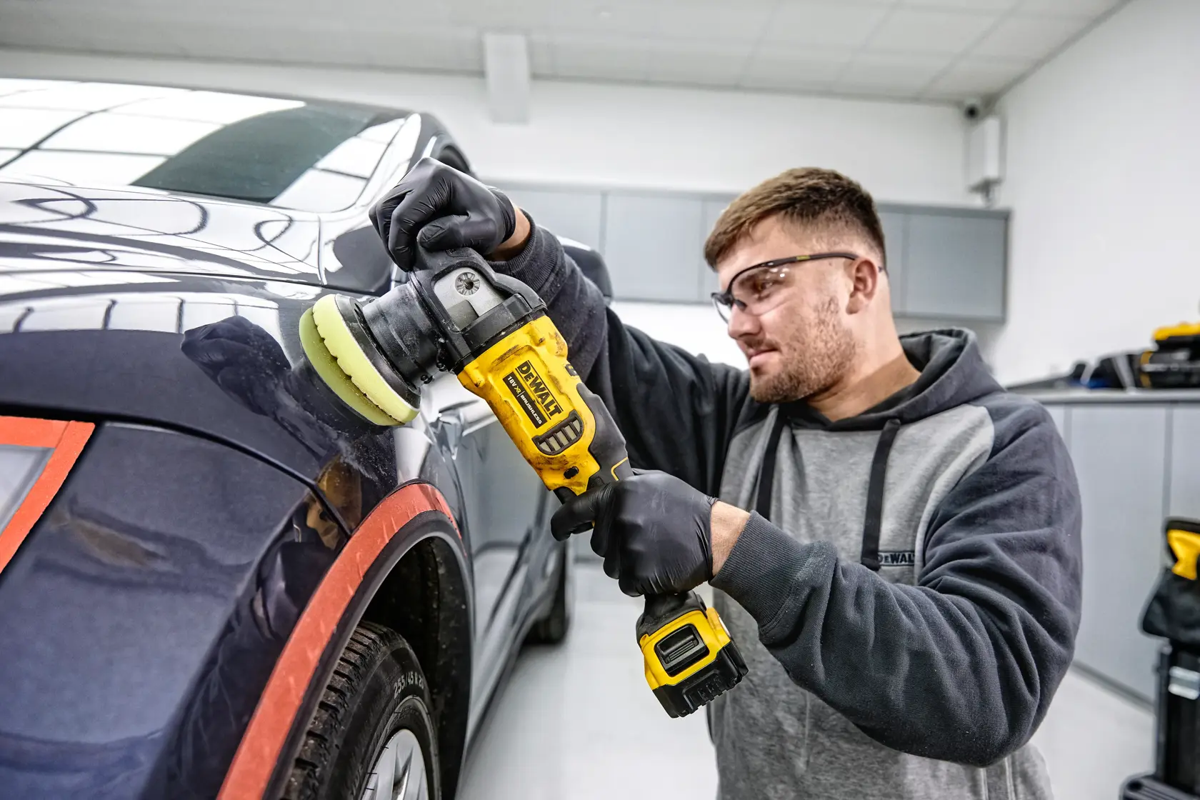 Person uses DEWALT cordless polisher DCM848P2 to polish a car in a workshop. Face is blurred for privacy.