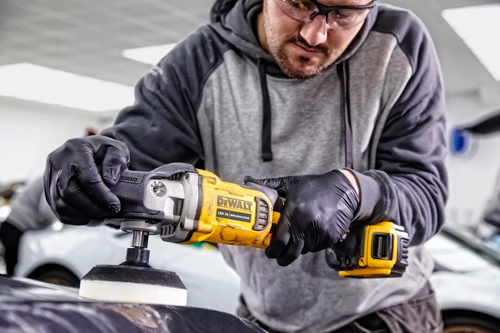 Person using a DEWALT DCM849P2 cordless polisher to work on a car surface in a garage, wearing gloves.