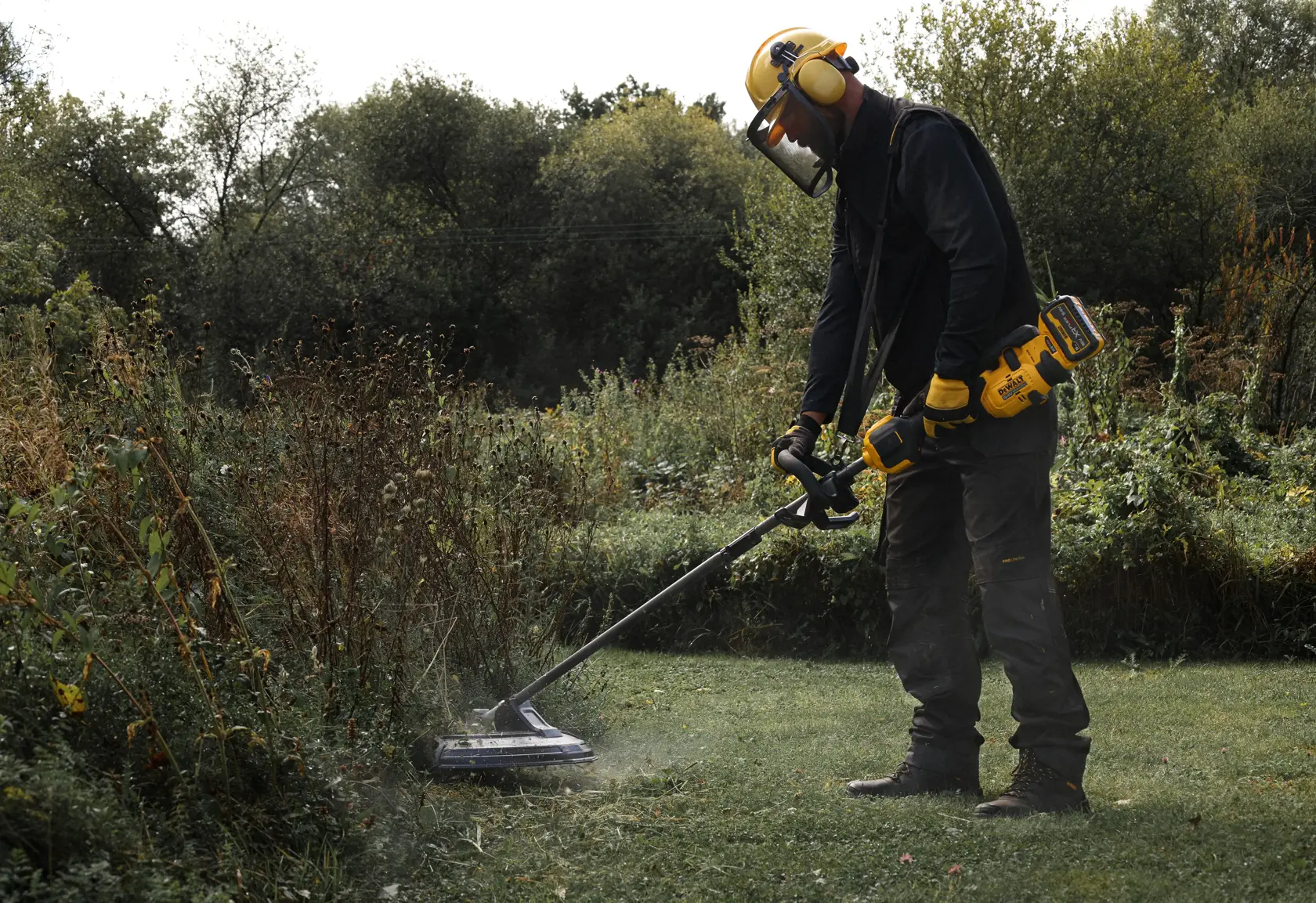 Person using DEWALT grass trimmer DCMASST1N, DCZ65713X1-XE, DCMASST1N-XJ outdoors. Includes safety gear.