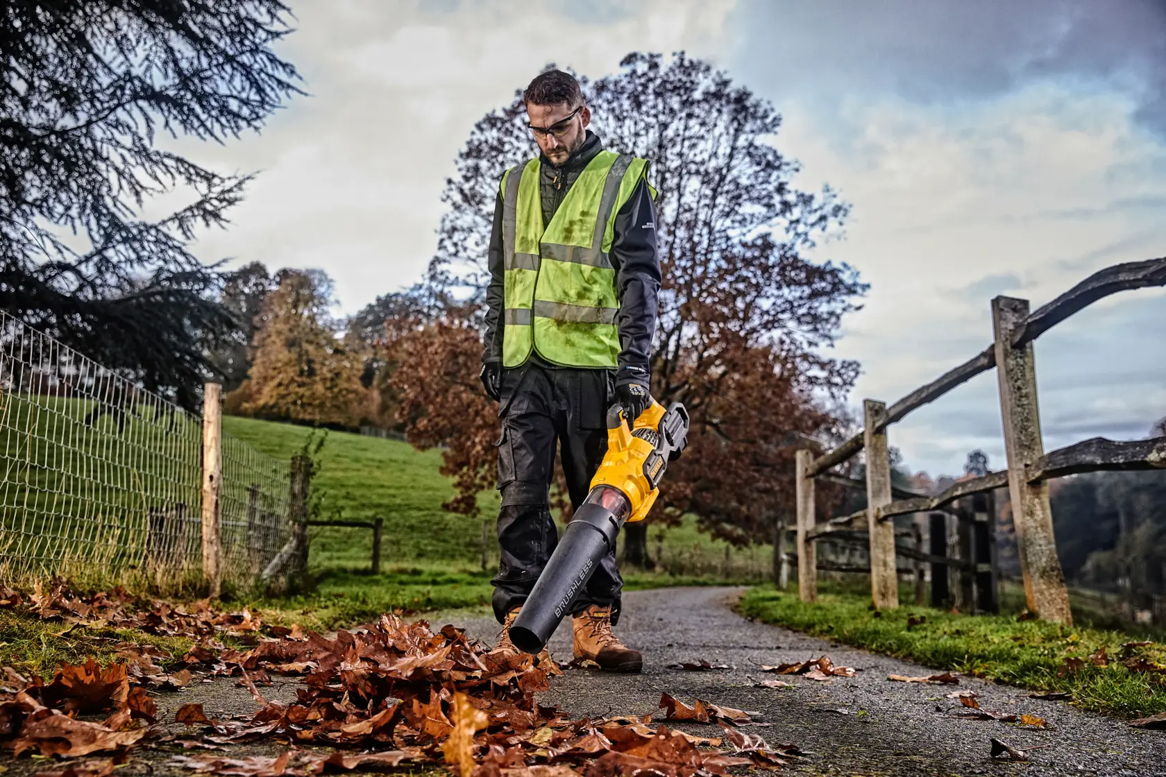 Person uses a DEWALT leaf blower on a rural path, wearing a safety vest, with autumn leaves on the ground.