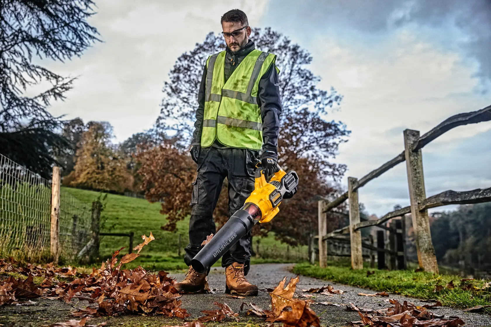 Person using a DEWALT leaf blower outdoors, wearing a safety vest and gloves, clearing leaves from a pathway.