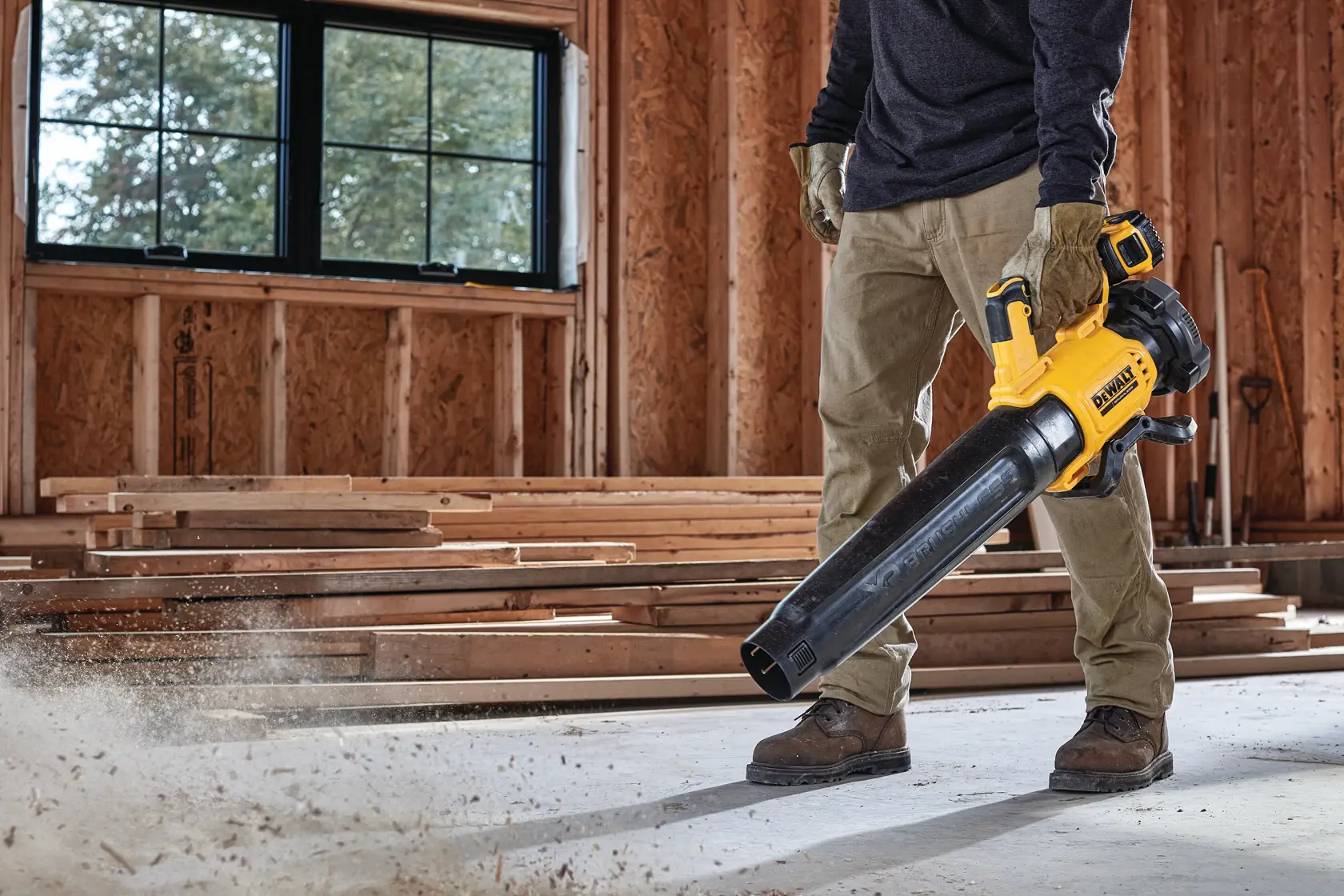Person using a DEWALT cordless blower to clear dust in a wood-framed room under construction.