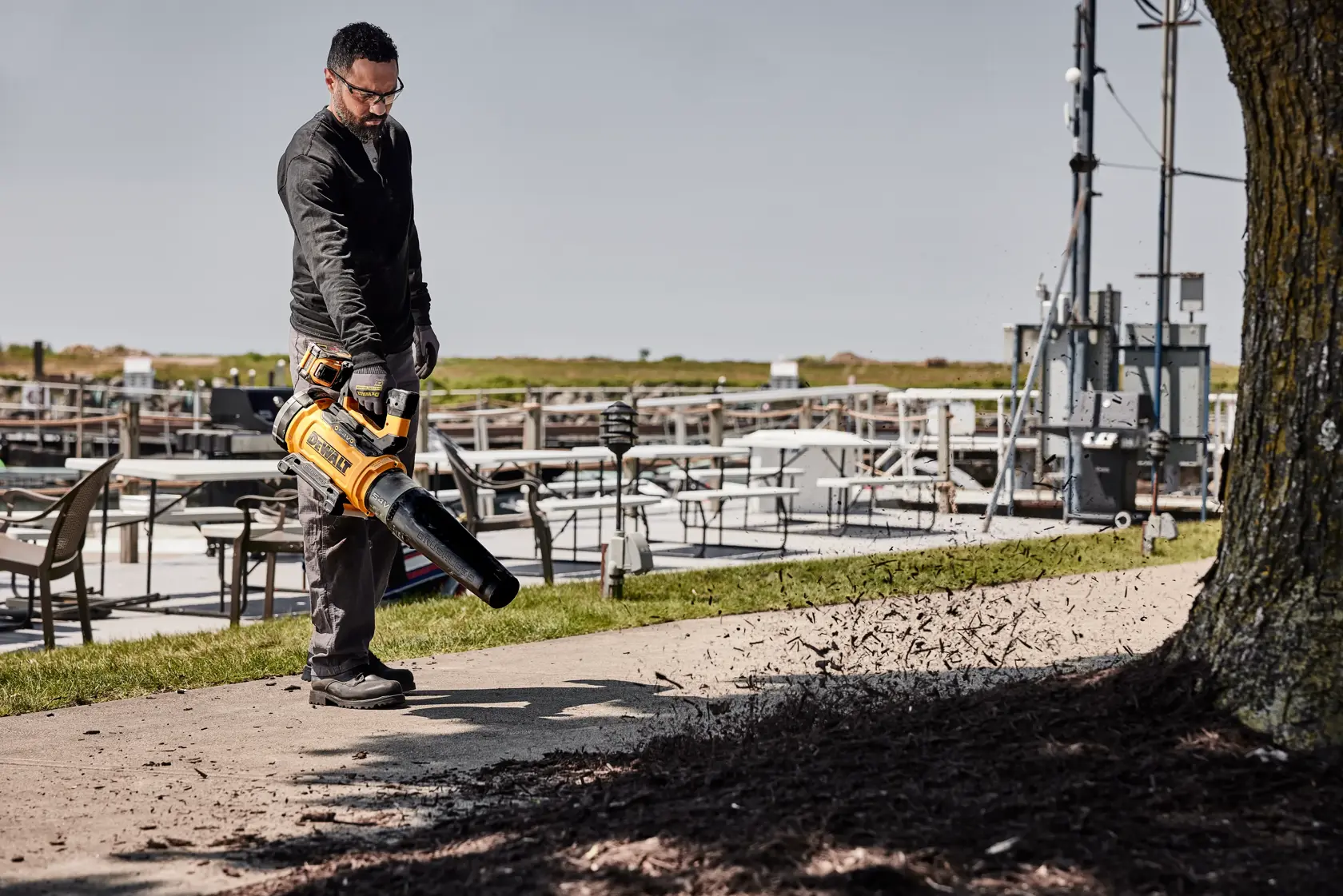 Person using a DEWALT cordless blower outdoors to clear debris from a sidewalk near chairs and tables.
