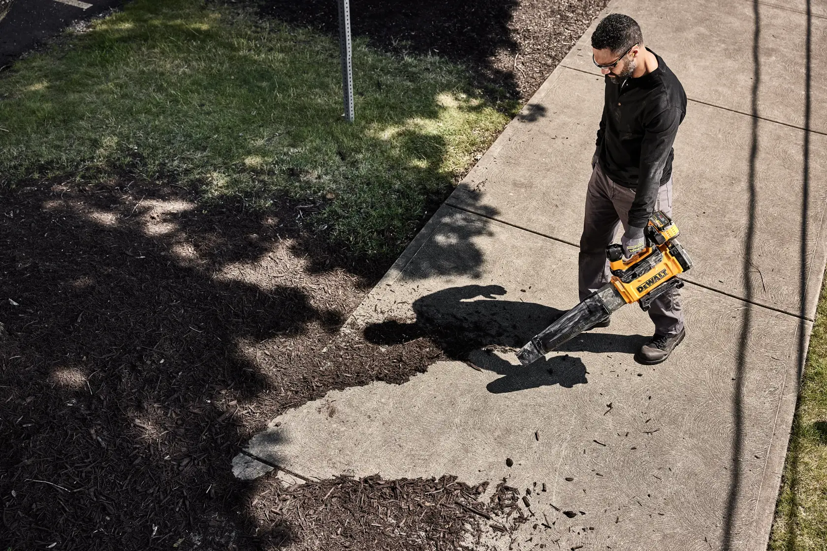 Person uses a DEWALT blower DCMBL777N to clear debris from a sidewalk outdoors.