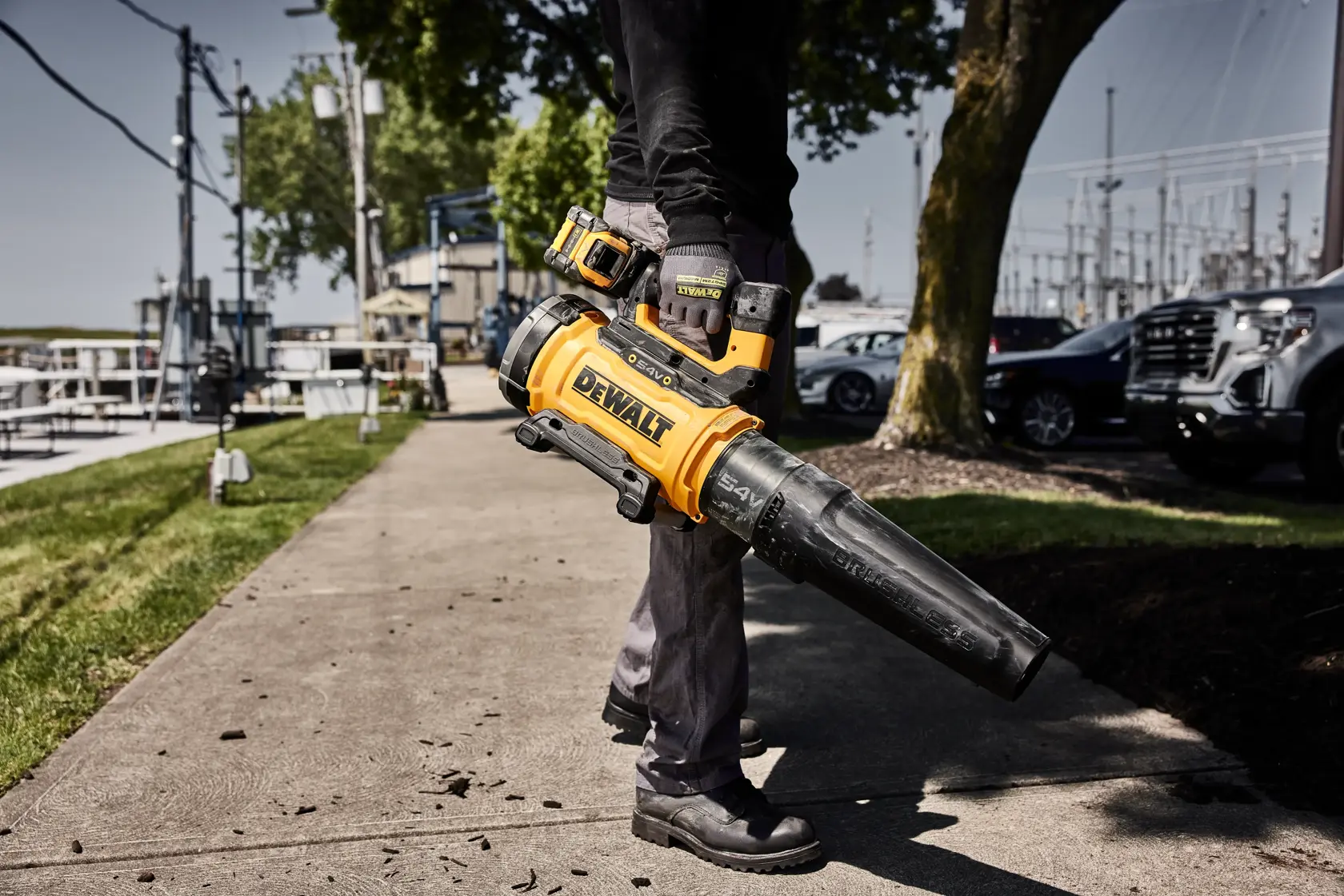 Person holding DEWALT cordless leaf blower while cleaning walkway outdoors near grass and parked cars.