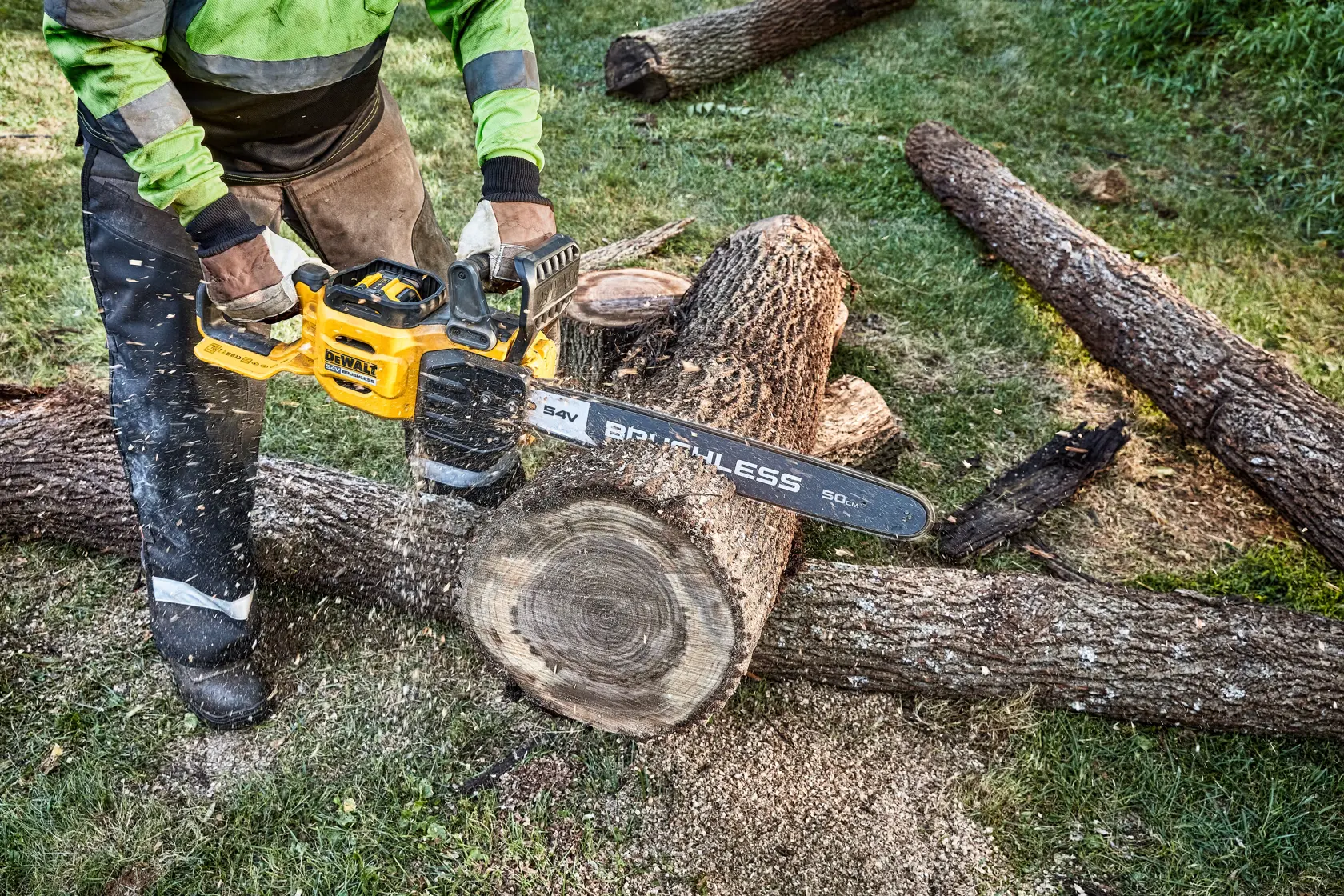 A person using a DEWALT cordless chainsaw to cut a large log outdoors, with wood chips and sawdust visible around the area.