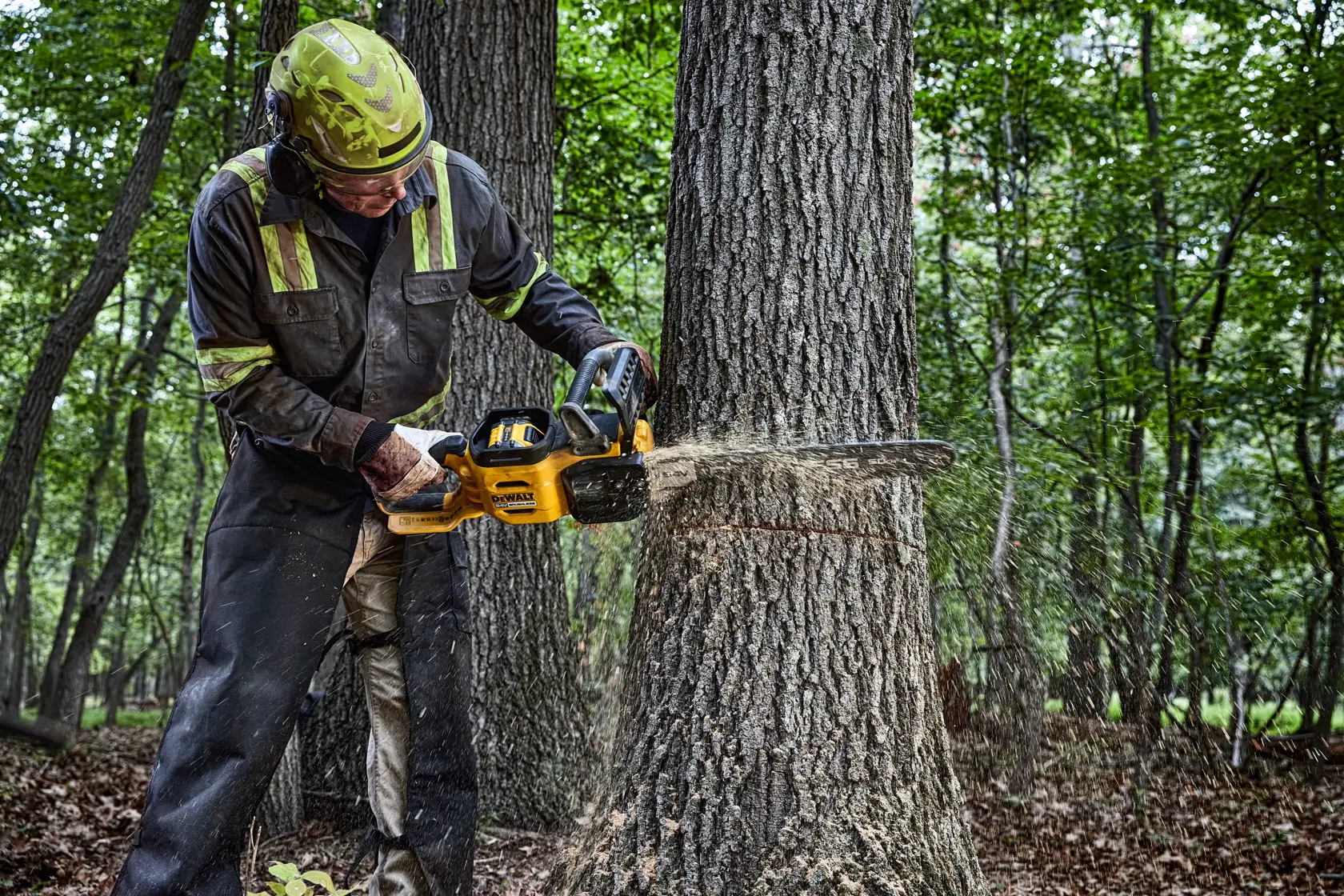 Una persona con indumenti protettivi utilizza una motosega gialla DEWALT per tagliare il tronco di un albero in un ambiente boschivo. La segatura è visibile durante il taglio del legno con la motosega.