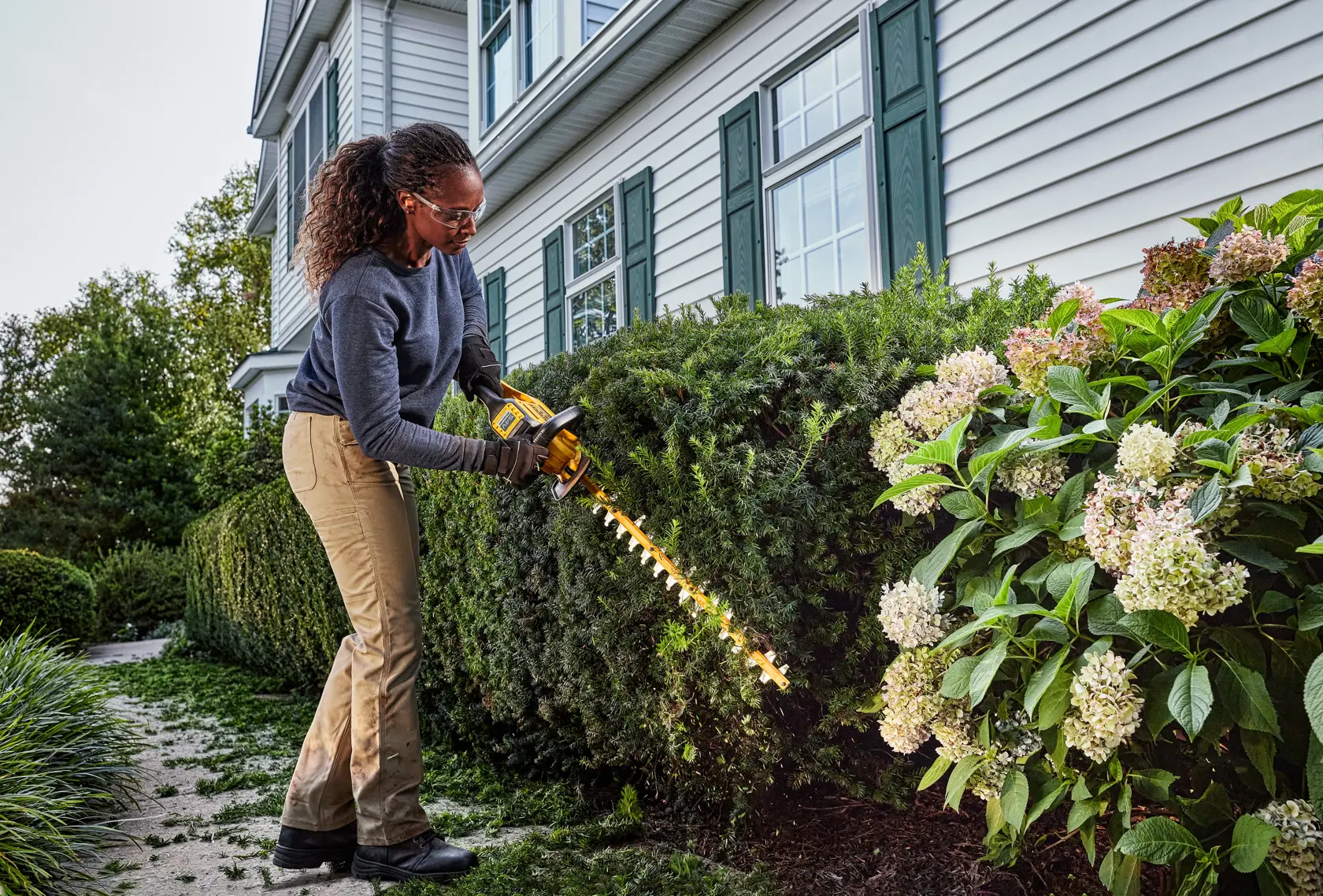 Persona usa un tagliasiepi a batteria DEWALT per tagliare siepi nel giardino di una casa.