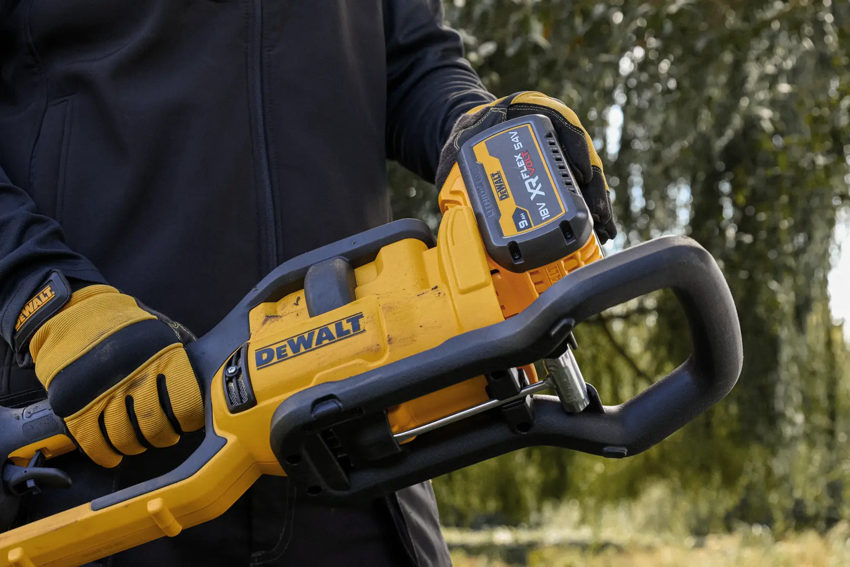 Gloved hands holding a DEWALT cordless chainsaw outdoors with a visible XR FLEXVOLT 18V battery.