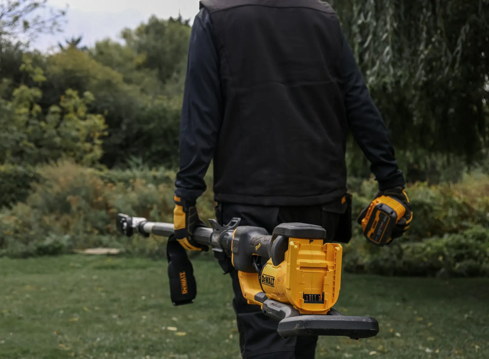 Person holding a yellow DEWALT power pruner outdoors, wearing gloves and workwear in a garden setting.
