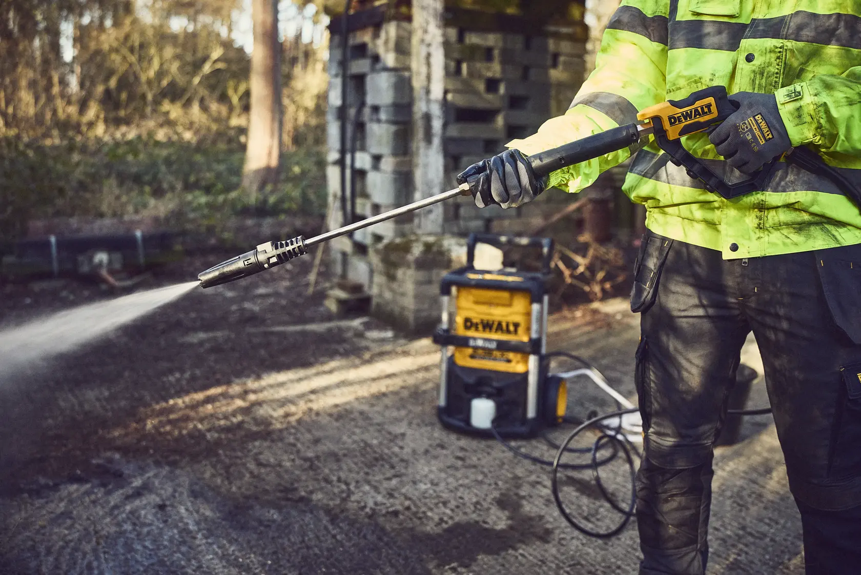 Person uses a DEWALT pressure washer outdoors on a paved surface beside a shed, wearing safety gear.