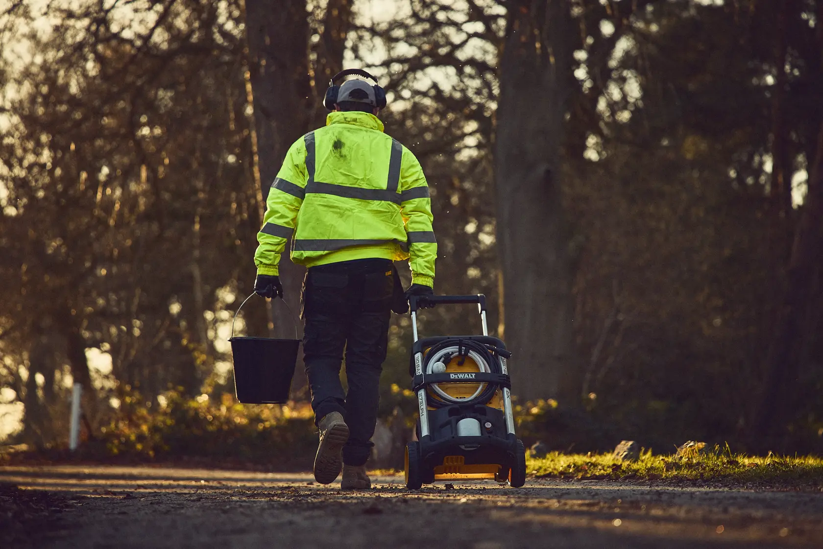 Person in safety gear pulls a DEWALT power washer and carries a bucket on a rural path at sunset.