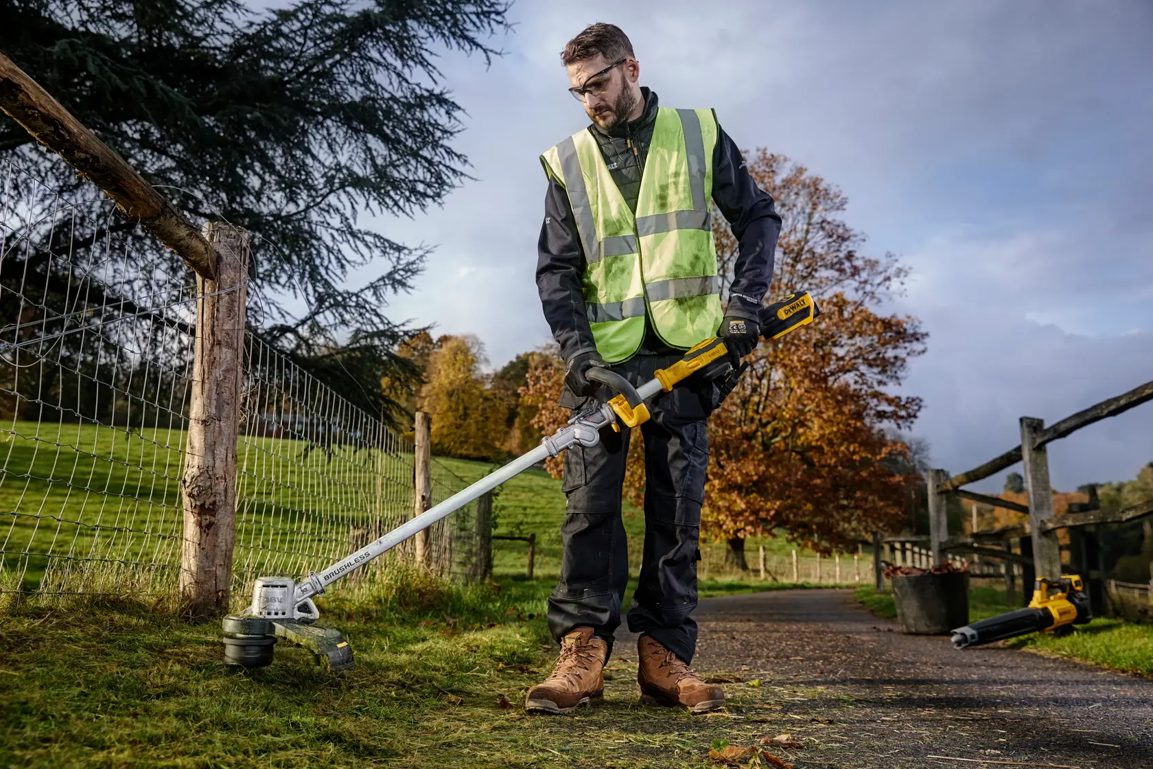 Person uses DEWALT grass trimmer outdoors while wearing a reflective vest and work boots by a fence.