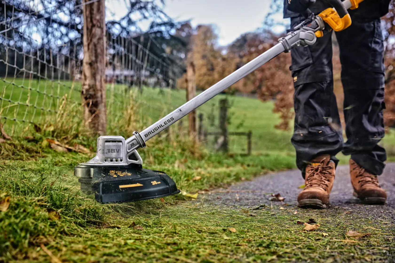 Persona taglia l’erba con un decespugliatore DEWALT brushless a batteria vicino a una recinzione.