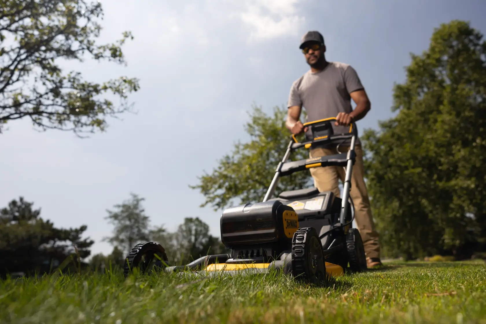 A person operating a DEWALT cordless lawn mower on a grassy lawn outdoors. The image shows the mower from a low angle, with blurred trees and sky in the background.