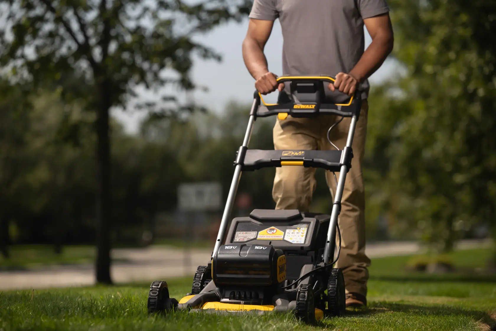 A person is pushing a DEWALT cordless electric lawn mower on green grass outdoors, with trees and a road visible in the background.