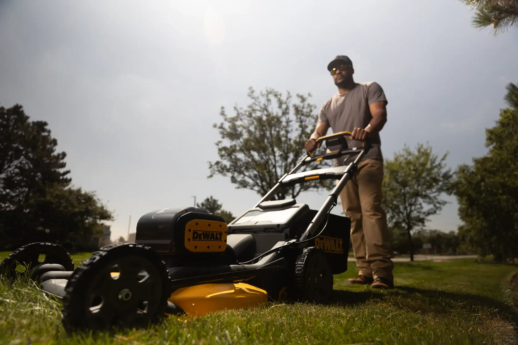 A person is using a DEWALT cordless lawn mower on a grassy area outdoors. The mower is black and yellow and the person’s face is blurred for privacy.